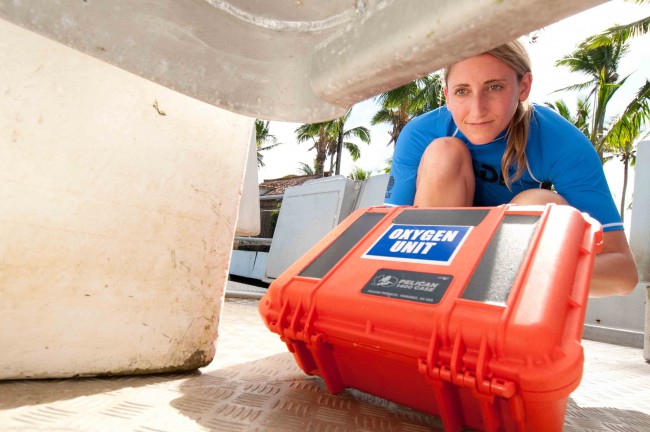 A diver retrieves an orange dry box with emergency oxygen equipment from under a bench