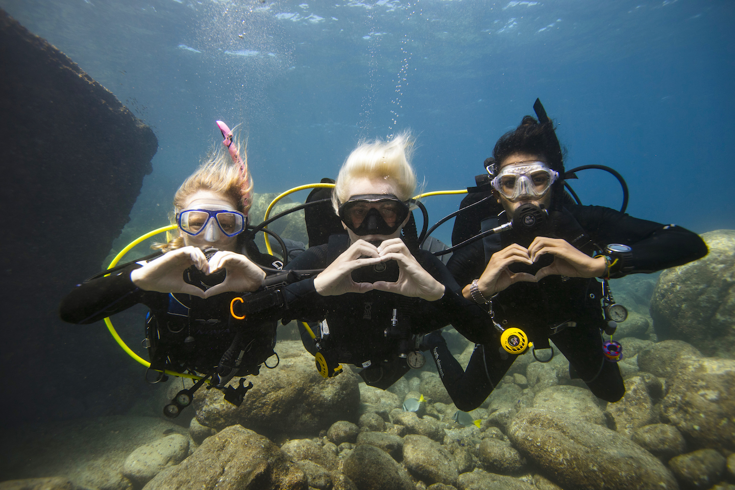 Three divers underwater making heart shapes with their hands, who might find love after sharing their best scuba diving jokes
