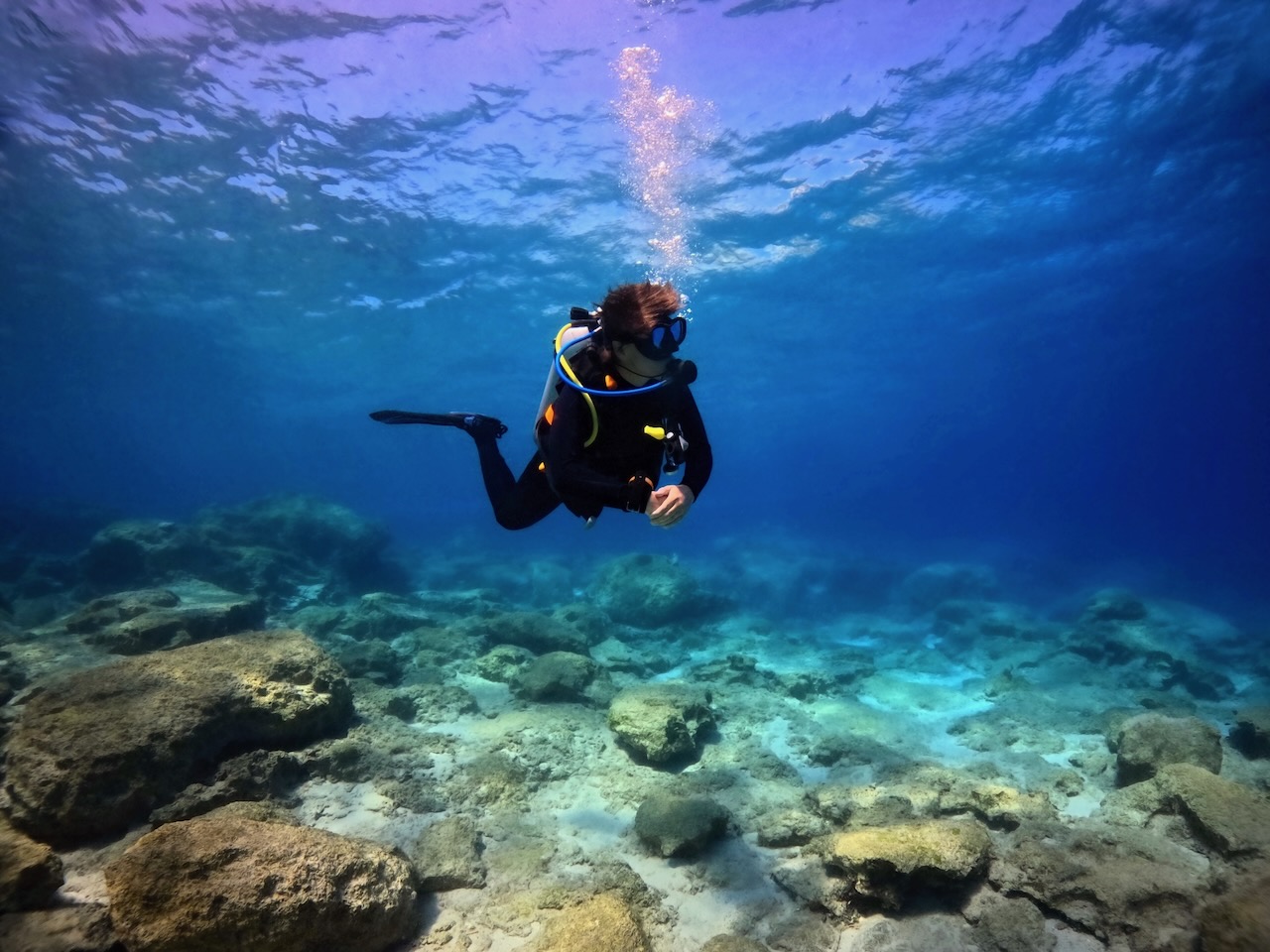 Scuba diver girl exploring shallow rocky seascape in the vivid blue clear sea. Underwater photography from scuba diving. Landscape in the ocean, travel photo. Stones and swimming diver.