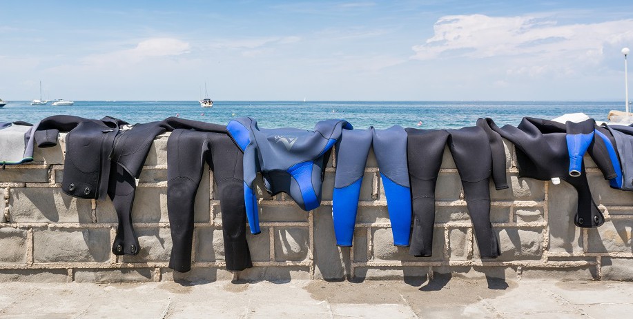 Several wetsuits hanging to dry on a wall near the water and which include shortie and full-length styles for scuba diving