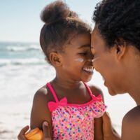 Mother embracing daughter with sunscreen on face at beach.