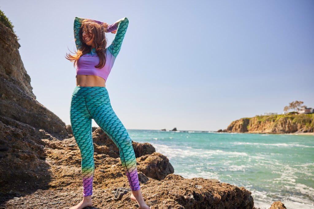 padi gear mermaid collection - a woman stretches on the seashore wearing cape cali leggings and padi rashguards