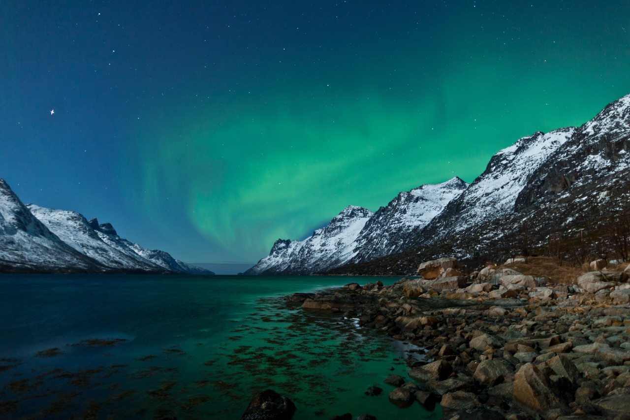 A topside shot of a tranquil Norweigan fjord at night, featuring ice-capped mountains and the northern lights across the sky
