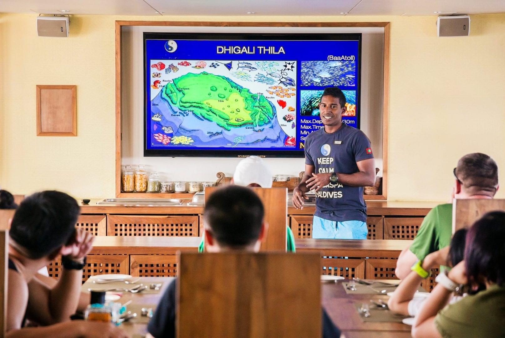 A PADI dive guide giving a dive site briefing about safety and marine life while on board a dive boat in the Maldives