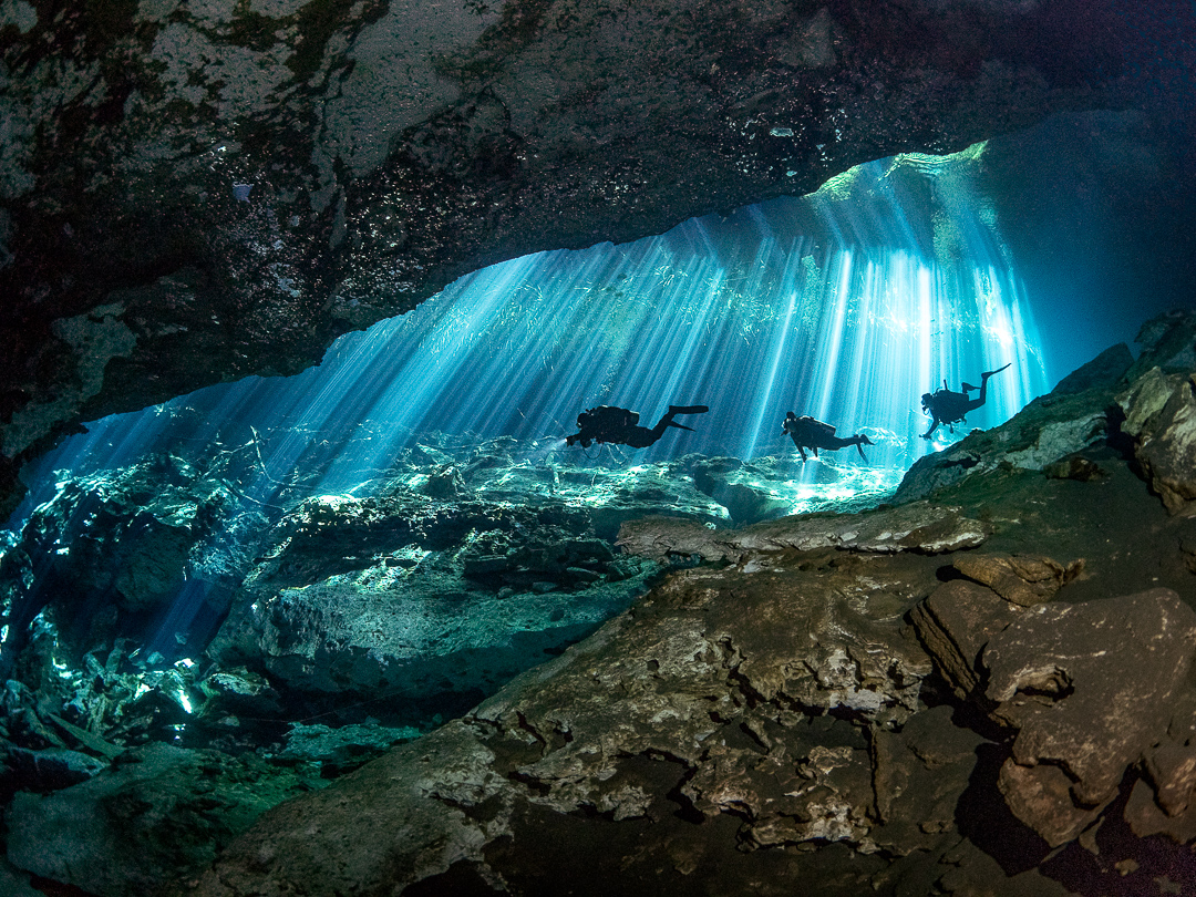 Three scuba divers swimming through a cenote in Mexico, where sunlight creates an ethereal effect that engages the senses