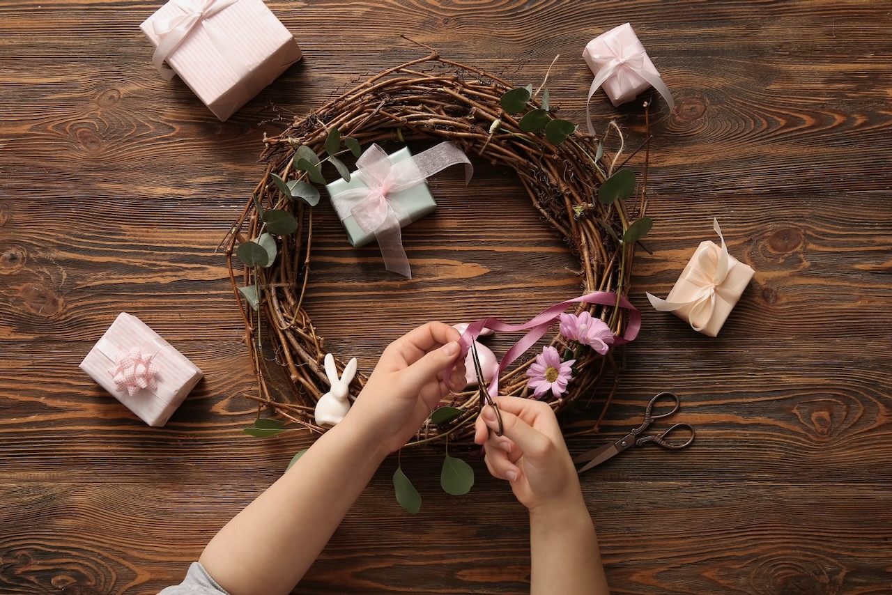 Woman making Easter wreath with flowers, rabbits and plant branches on brown wooden background