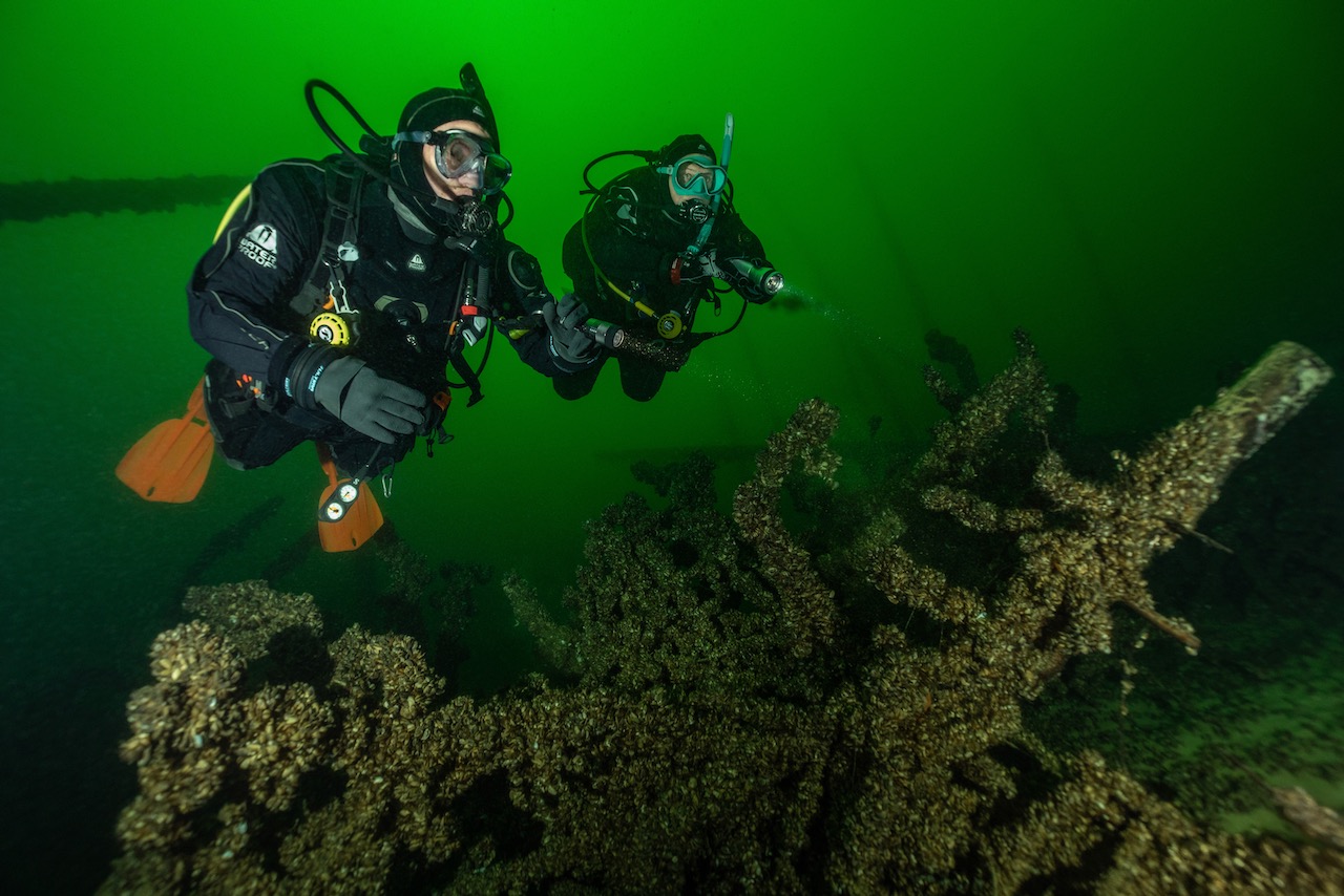 two scuba divers explore the green waters of the DACH region of Europe. They are wearing dry suits.
