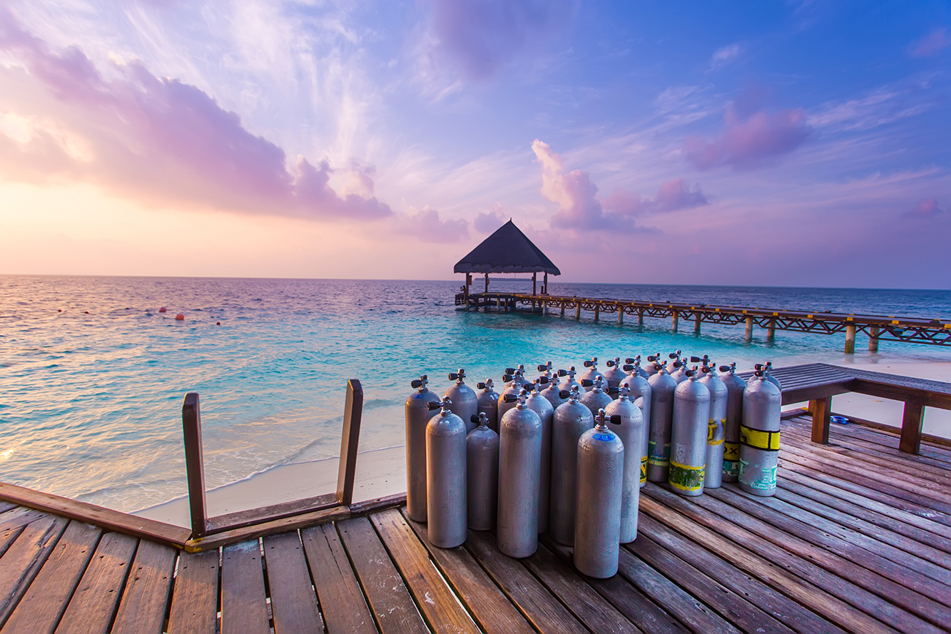 A collection of scuba diving tanks stood on a deck next to the water's edge during a beautiful pink sunset in the Maldives