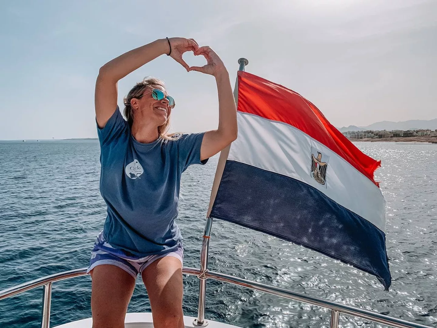 A woman makes a heart symbol with her hands while sitting on the edge of a boat next to an Egyptian flag