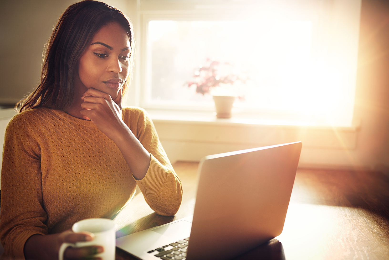 A person sits in front of a laptop holding a mug and thinking