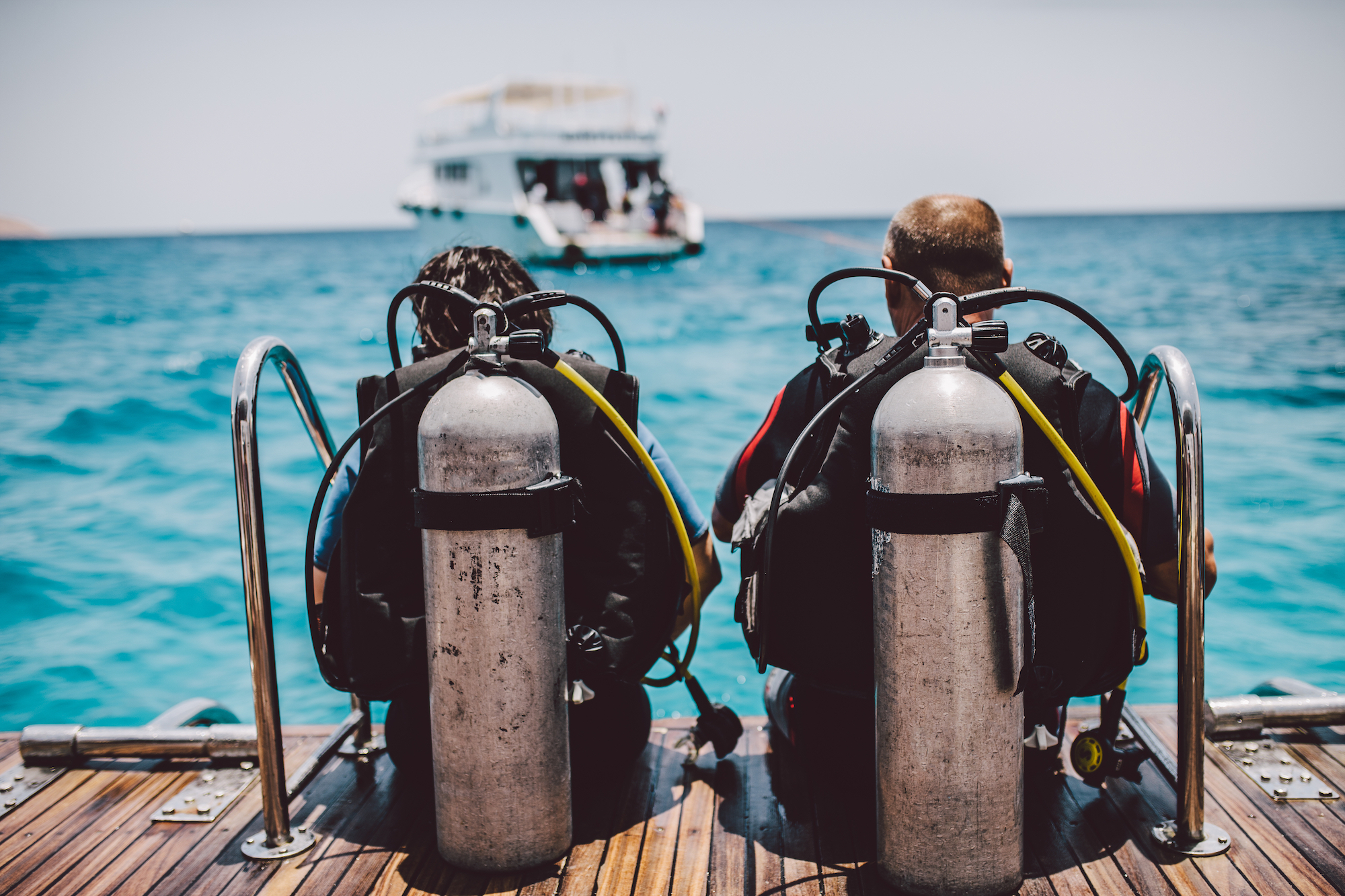 Two divers sitting on the deck of a boat, preparing to go scuba diving, which is lower risk than many other popular sports