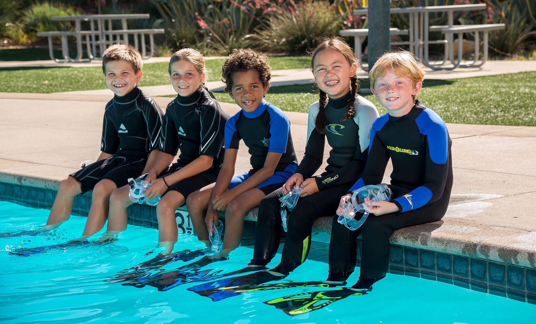 A group of five young scuba divers sitting on the side of a swimming pool before starting their Bubblemaker lesson for kids