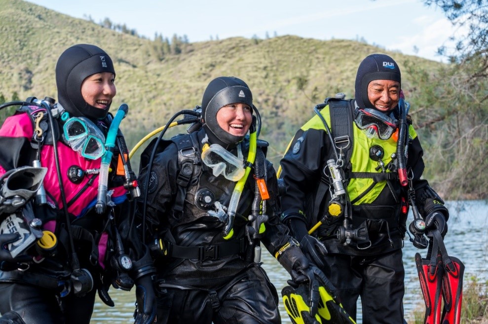 Three divers in dry suits and smiling after a cold water dive which answers the question 'are drysuits warmer than wetsuits?'