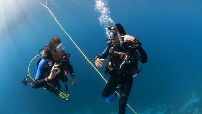 Two divers underwater. One signals to the other they are experiencing ear problems.
