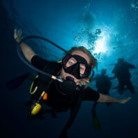 a scuba diver looking down at the camera while other divers hover above