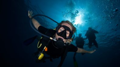 a scuba diver looking down at the camera while other divers hover above