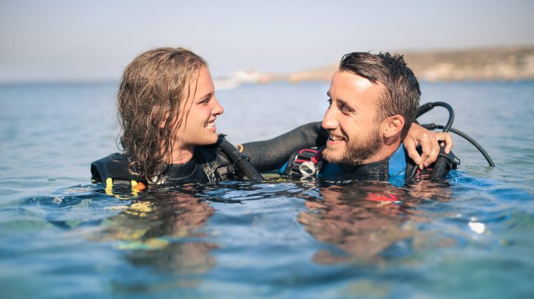 Two scuba divers float at the surface smiling at each other. The diver on the left has her arm around the other in a friendly gesture.