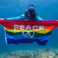 A scuba diver holds up a rainbow flag with the word peace on it.