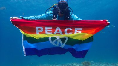A scuba diver holds up a rainbow flag with the word peace on it.