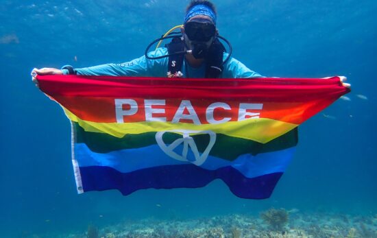 A scuba diver holds up a rainbow flag with the word peace on it.
