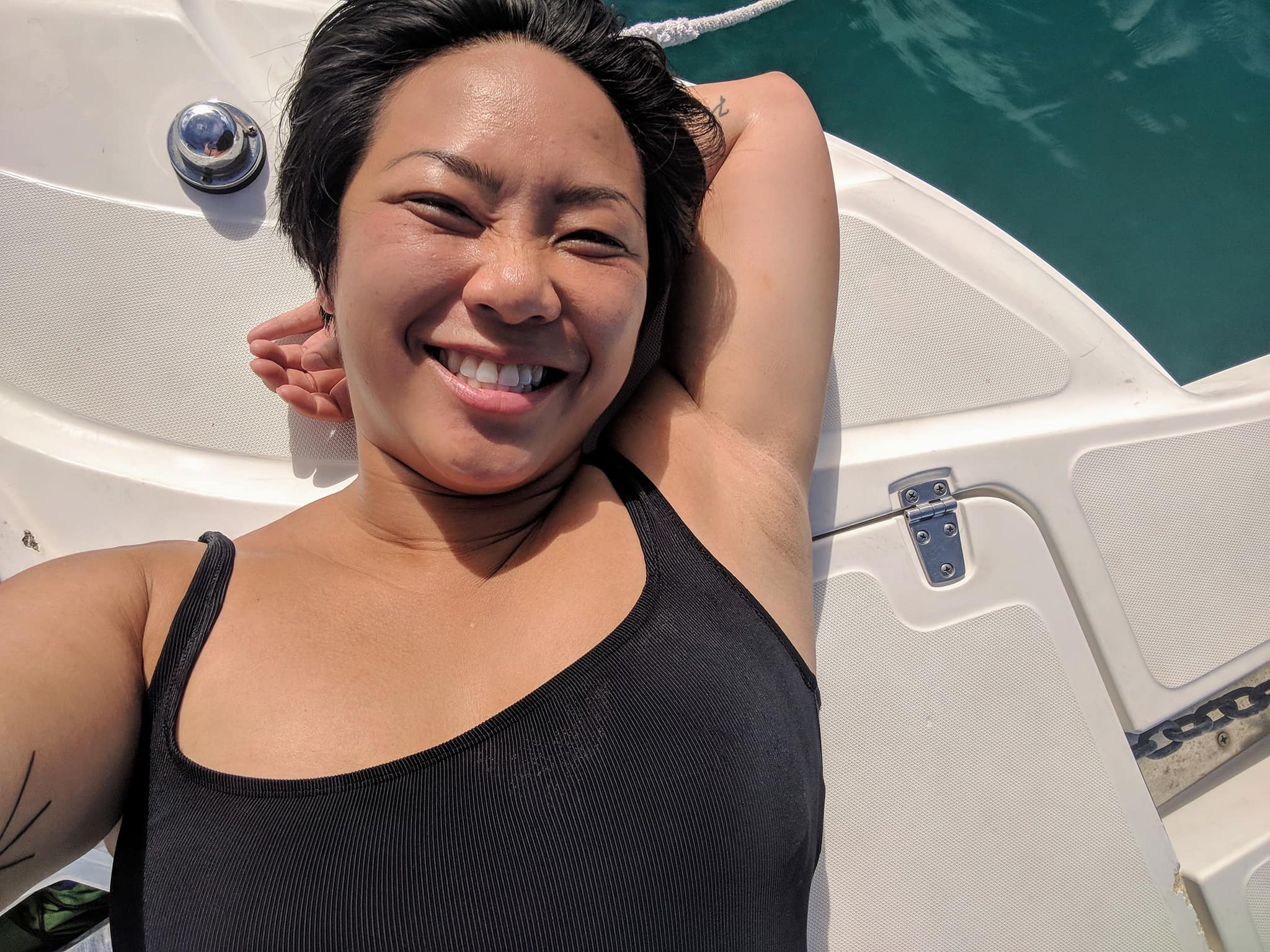 A woman in a black bathingsuit smiles whlie laying on the bow of a boat.