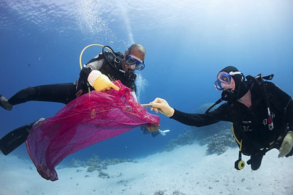 Two divers collect marine debris into a pink mesh bag.