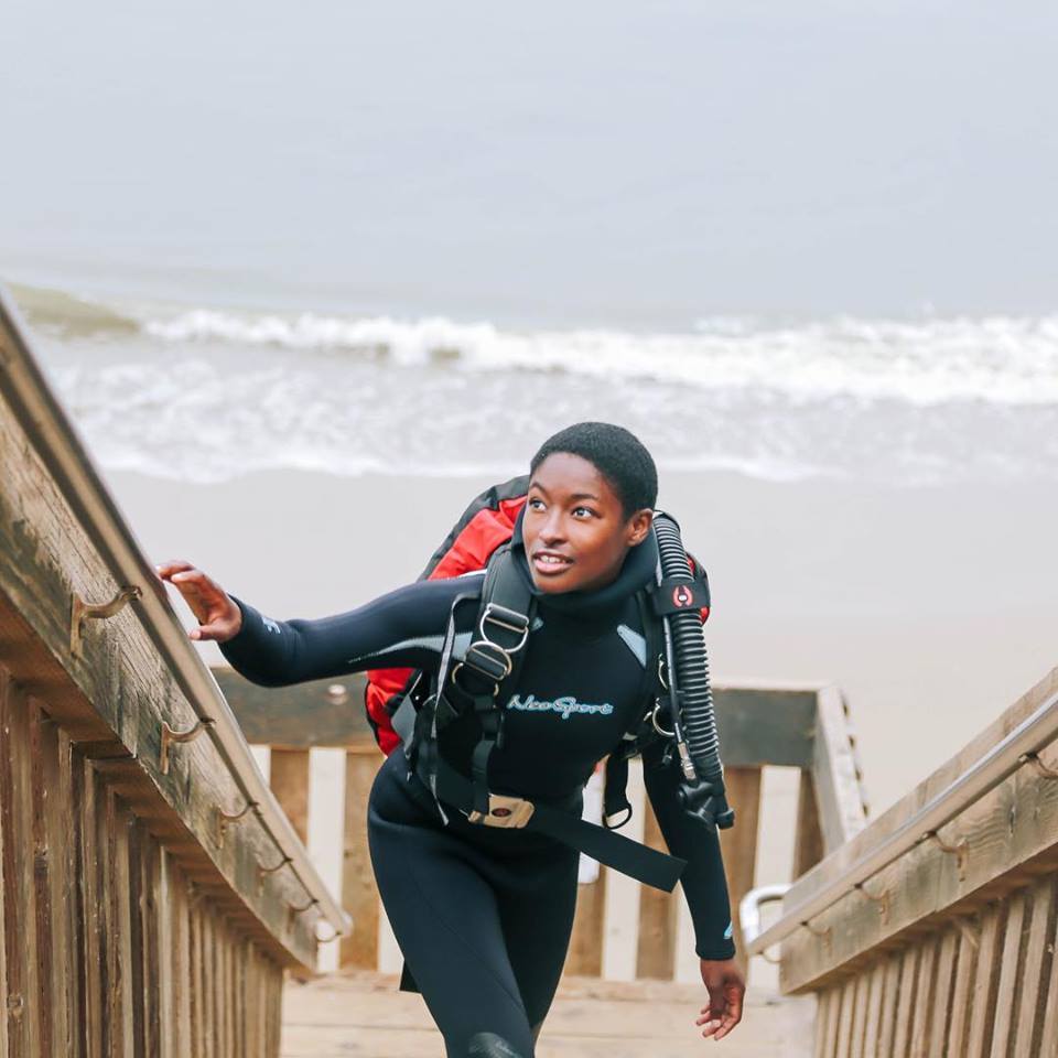 A Black woman diver in her gear climbs up steps from a beach