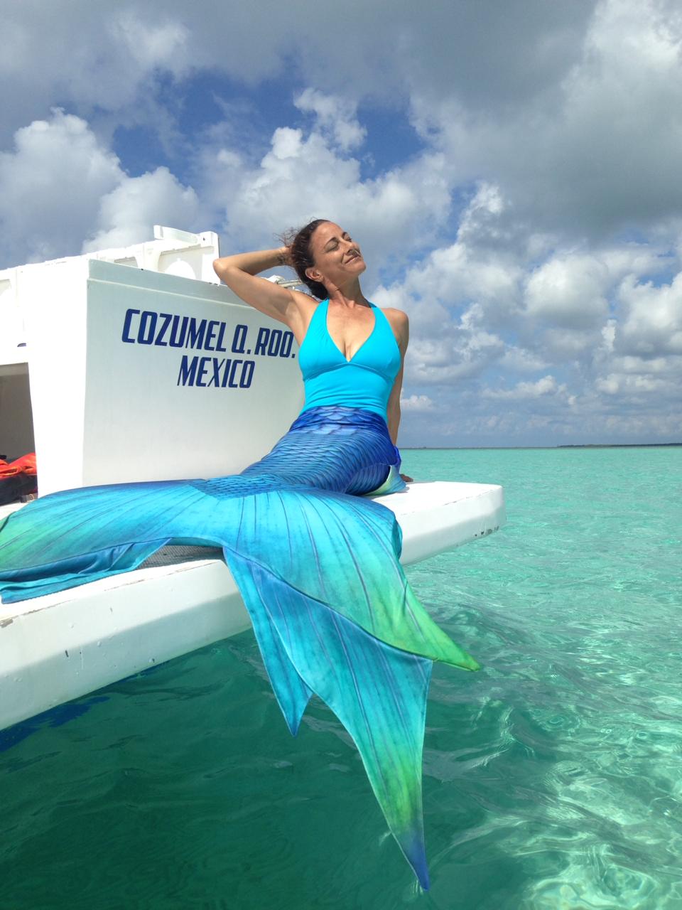 A woman in a blue and green mermaid tail lays across the side of a boat.