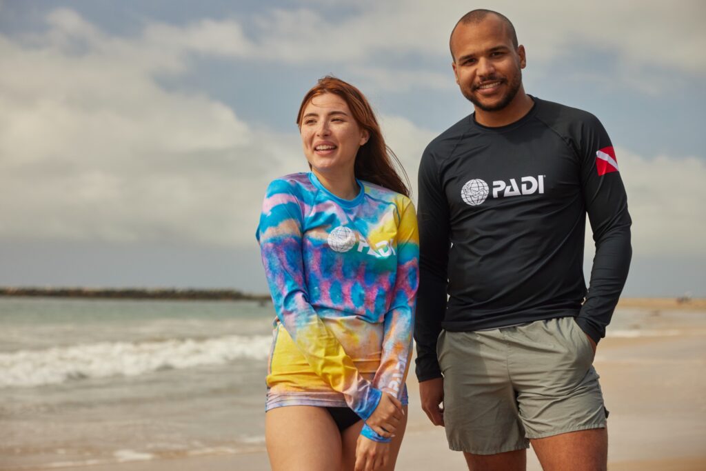 two people stand together smiling on the beach wearing PADI rashguards