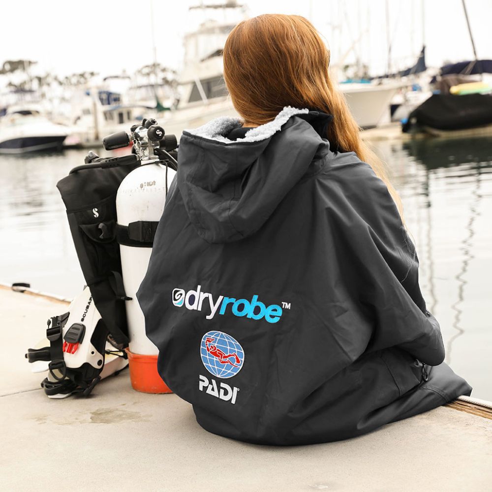 A girl sits in a marina next to dive gear. she's wearing a PADI branded dryrobe.