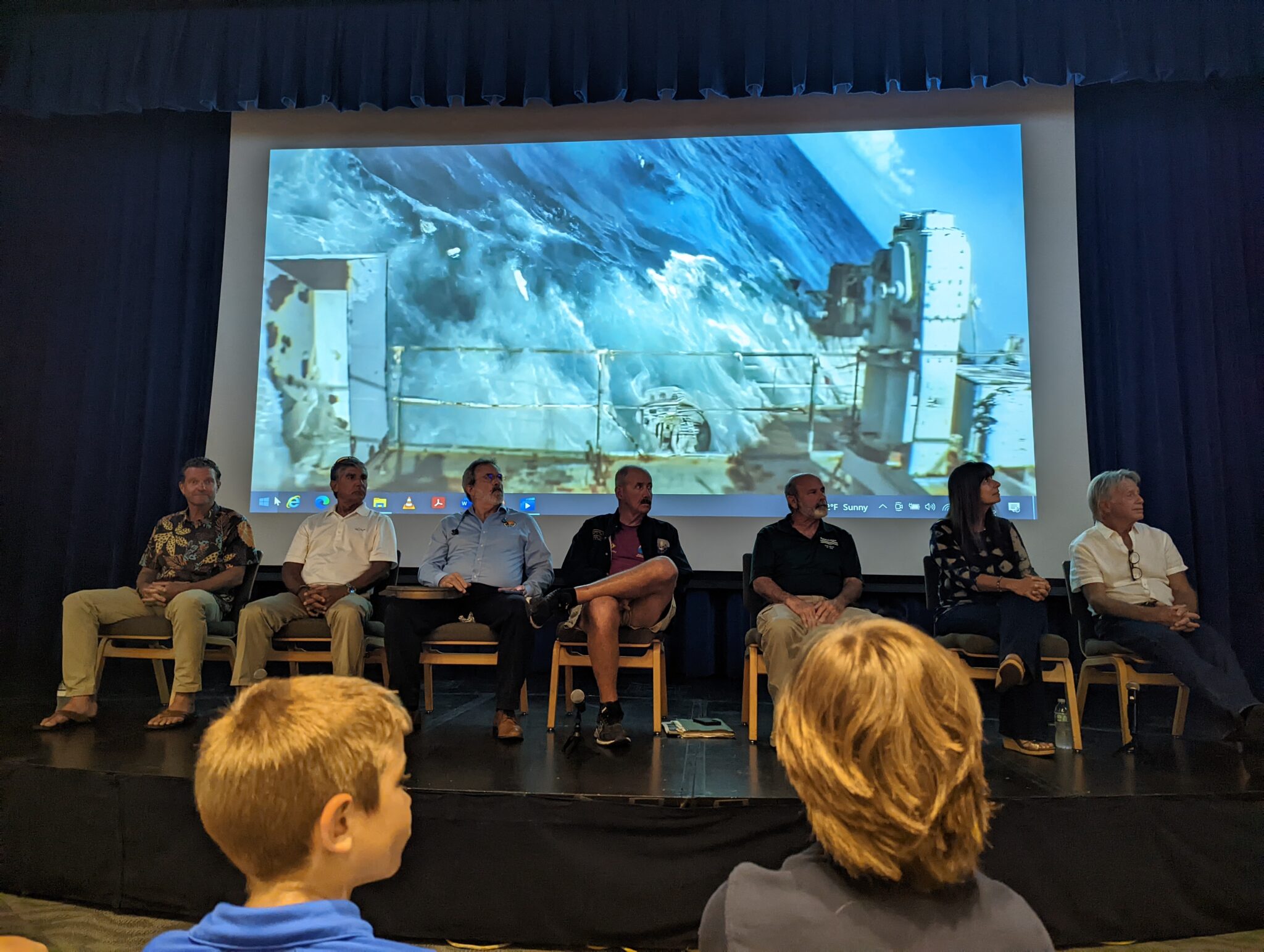 A panel of speakers sit in front of a movie showing a ship sinking