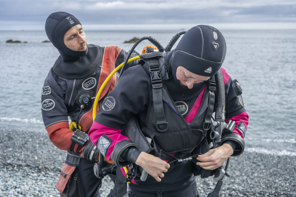 A diver helps their buddy. Both wear dry suits.