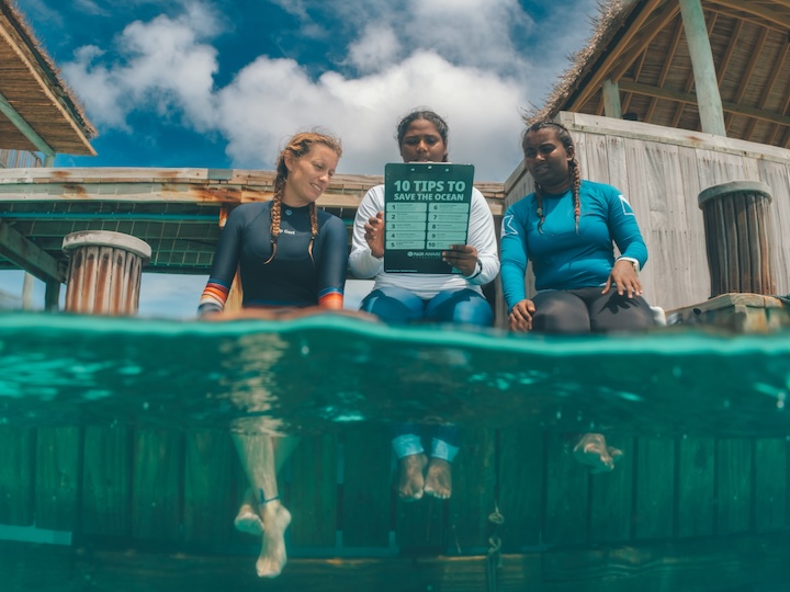 três mergulhadores seguram uma placa de conservação enquanto estão sentados na beira de uma piscina