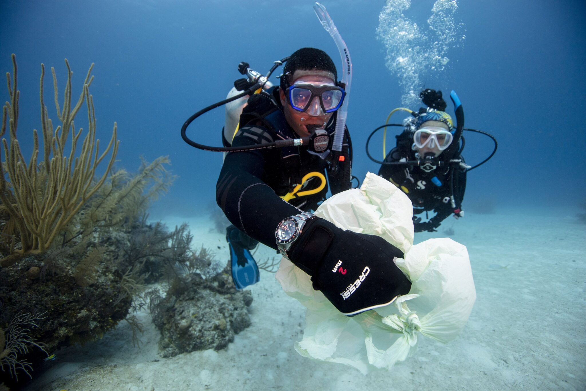 Twee duikers verzamelen afval onder water, één duiker houdt een plastic zak vast