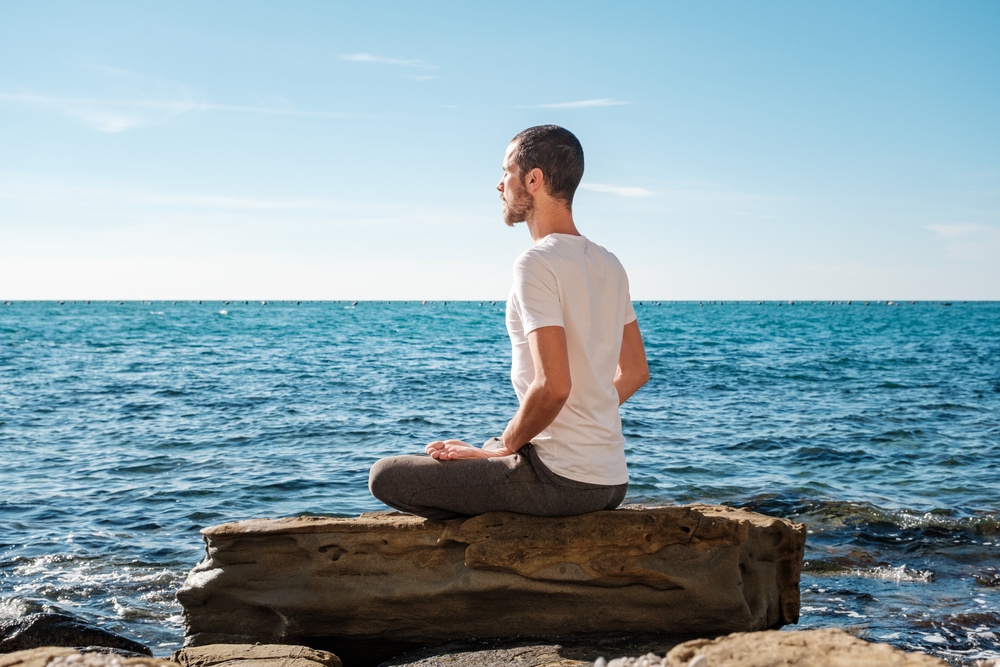 A mans sits cross legged on a rock by the ocean