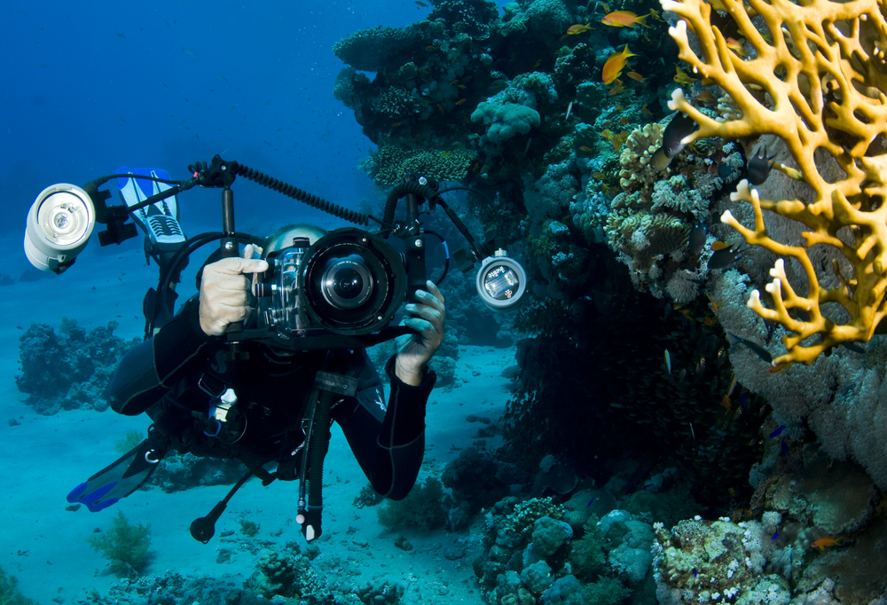 An underwater photographer with strobes extended takes a photo of something on the reef