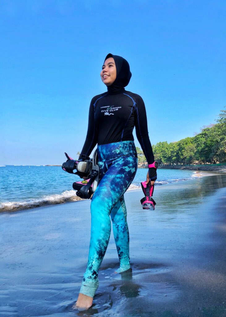 A Muslim woman diver stands at the shoreline, holding her mask and fins.