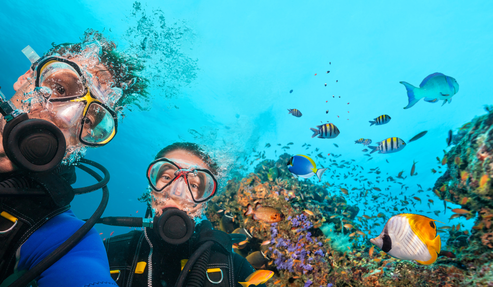 Two Master Scuba Divers smiling together while underwater against a backdrop of a colorful coral reef and tropical fish