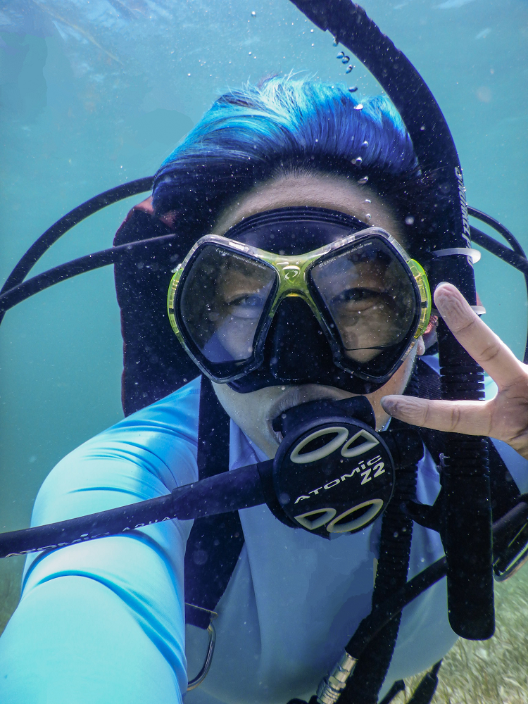 A diver with blue hair holds up a peace sign underwater while taking a selfie