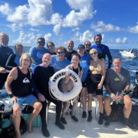 A group of 13 smiling scuba divers on a boat posing with a white life ring with the ocean in the background