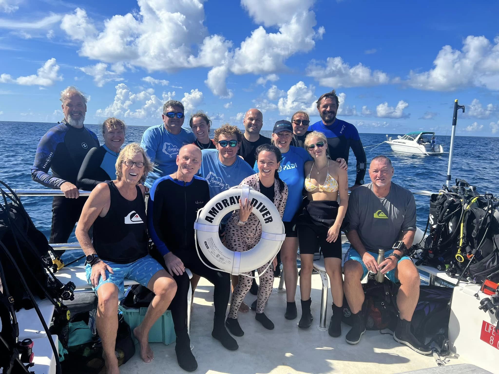A group of 13 smiling scuba divers on a boat posing with a white life ring with the ocean in the background