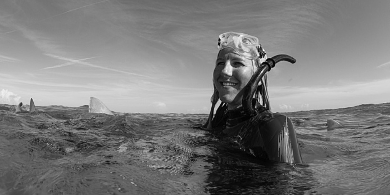 A freediver with her mask up smiles with shark fins in the background in a black and white photo.