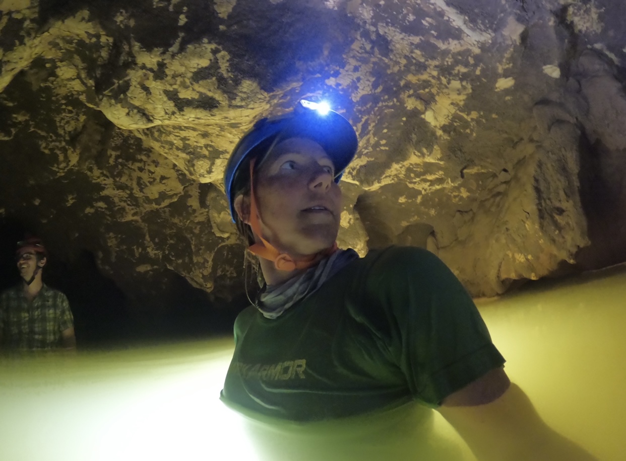 A woman walks chest-deep in water in a cave. She has a headlight on.