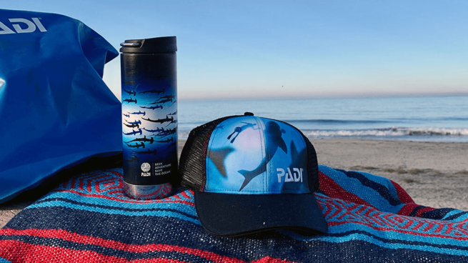 A PADI dry bag, thermos and hat sit on a woven blanket on the sand with the ocean in the background.