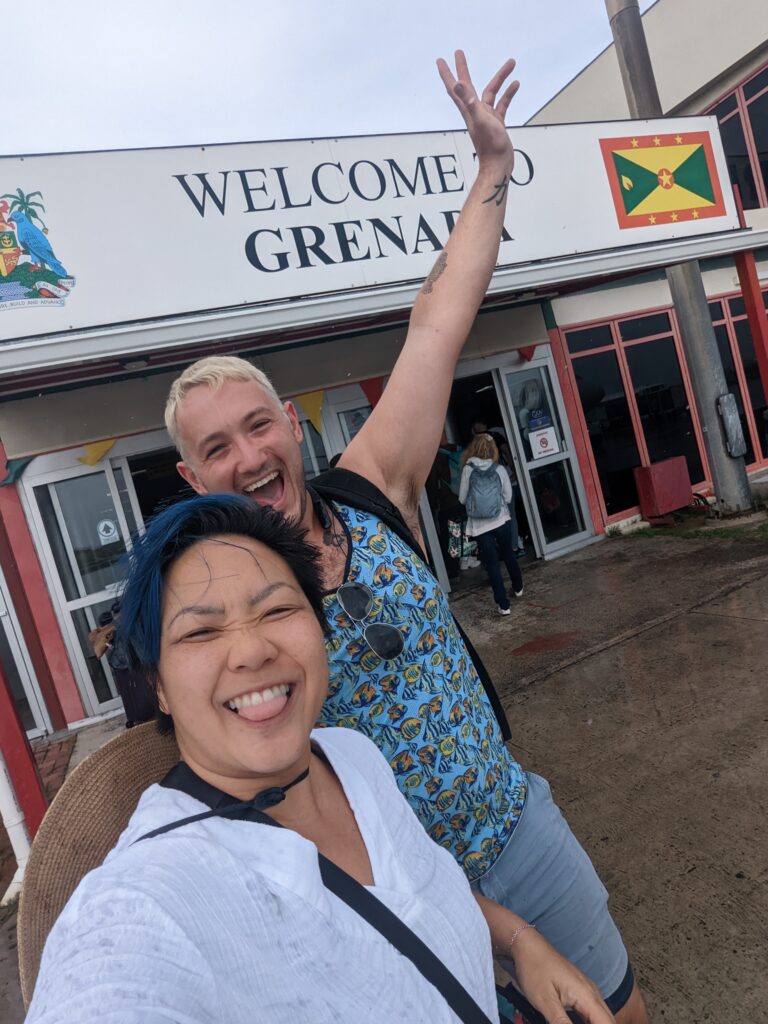 A woman and man excitedly smile in front of an airport sign that reads, "Welcome to Grenada." Dive Resorts are often easy to reach.