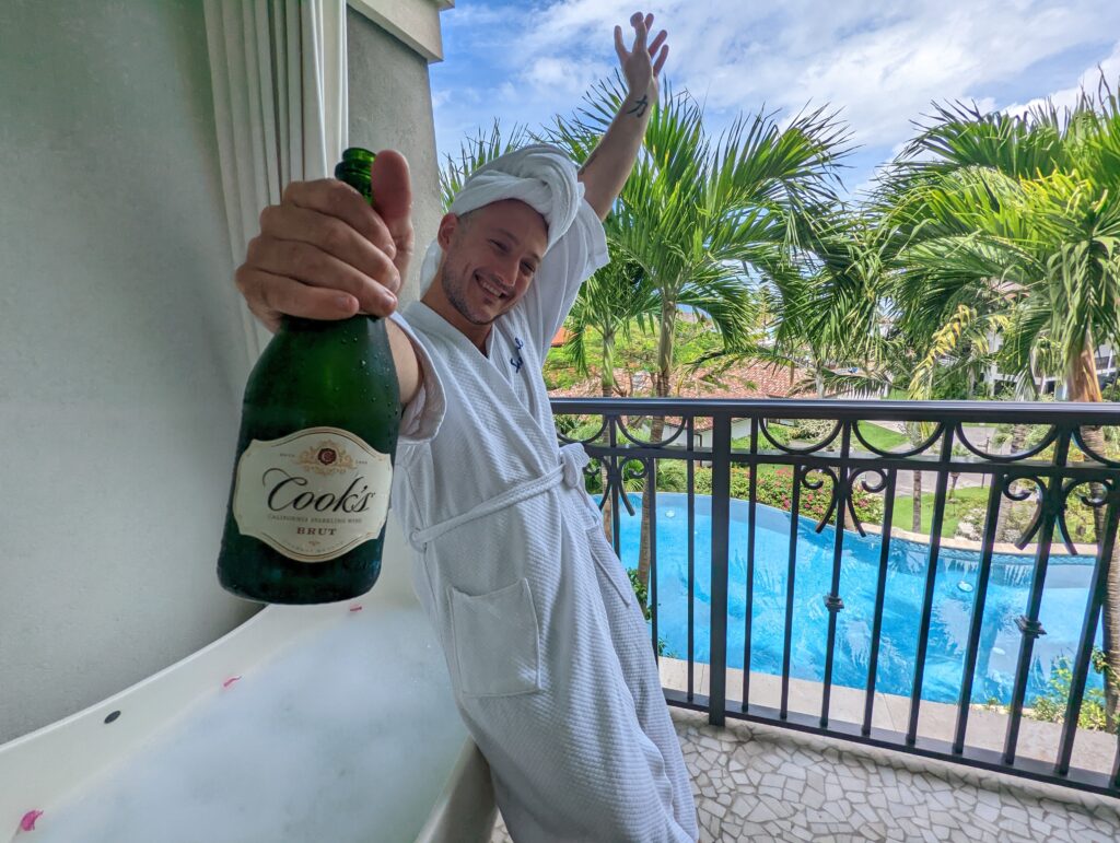 A man in a robe with a towel on his head leans on a bathtub full of bubbles. He holds a bottle of champagne in front of him and celebrates being at Sandals Resorts.