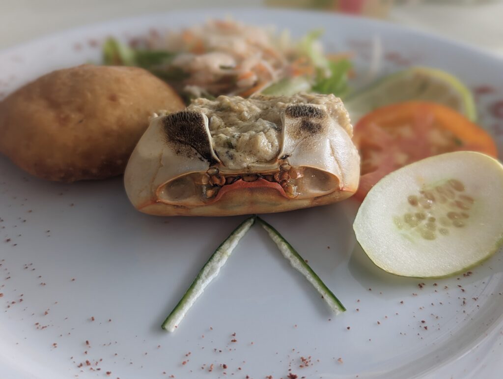 A crabshell overturned and stuffed with crab meat and other ingredients sits on a plate with other garnish and a piece of bread.