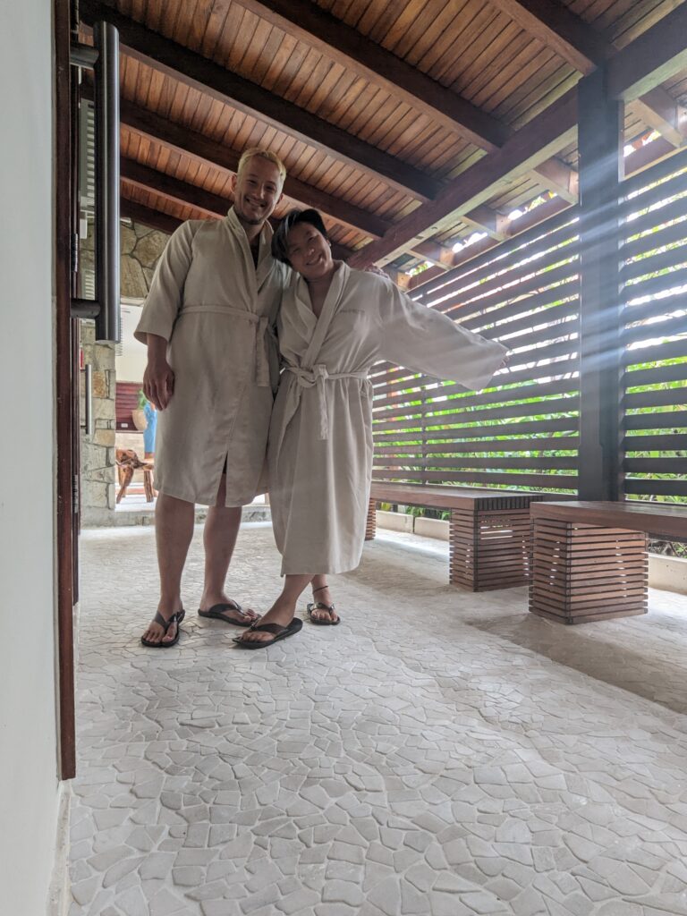 Two people in white robes pose in front of wooden slats at a spa at Sandals Resorts Grenada.