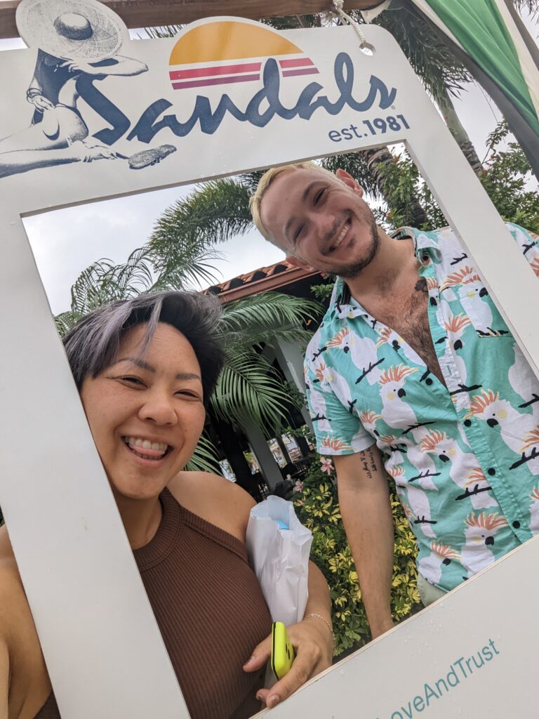 A woman and man stand behind a giant "polaroid" frame that says "Sandals"