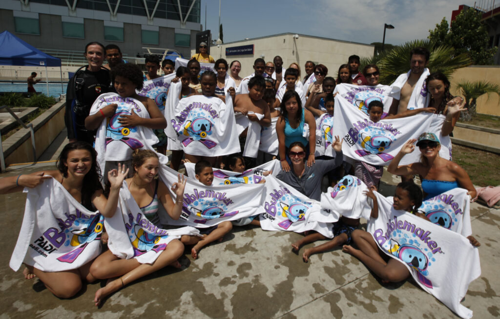 A group of kids hold up white towels that say "BubbleMakers" on them after learning to scuba dive.